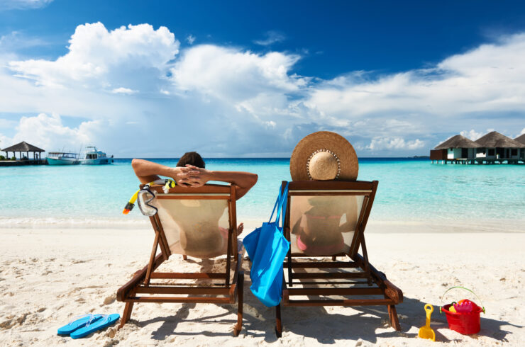 Two people are relaxing in beach chairs on a beach. They are looking out at the sea, clouds, and small vacation homes.