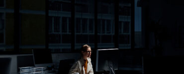 אישה צעירה יושבת ליד השולחן שלה במשרד חשוך בזמן משמרת הלילה שלה, על השולחן יש לה מחשב שמאיר על פניה