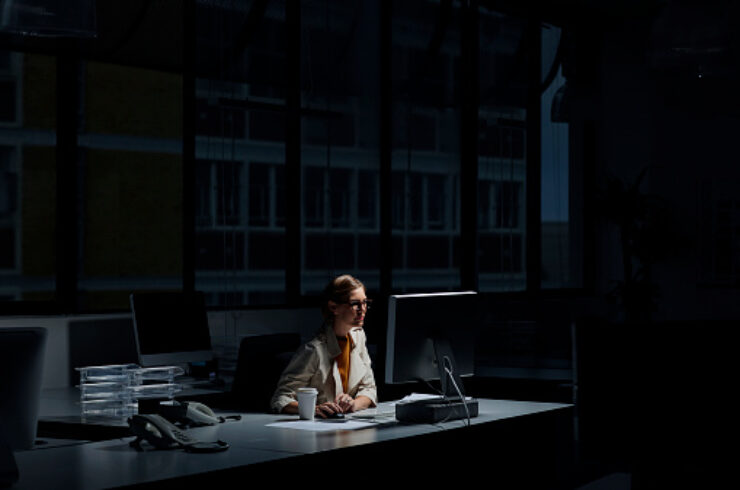 אישה צעירה יושבת ליד השולחן שלה במשרד חשוך בזמן משמרת הלילה שלה, על השולחן יש לה מחשב שמאיר על פניה