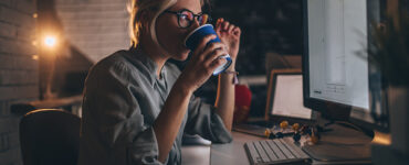 בתמונה אישה בלונדינית צעירה יושבת בלילה ליד השולחן שלה בעבודה ומול המחשב, עובדת שעות נוספות ושותה קפה כדי להשאר ערנית