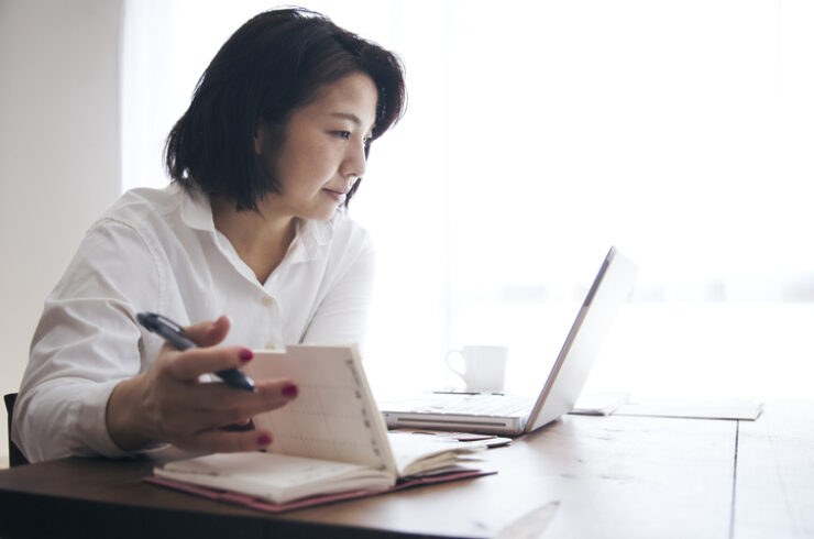 אישה יפנית בבגדי יומיום בודקת וכותבת את לוח הזמנים ביומנה על השולחן